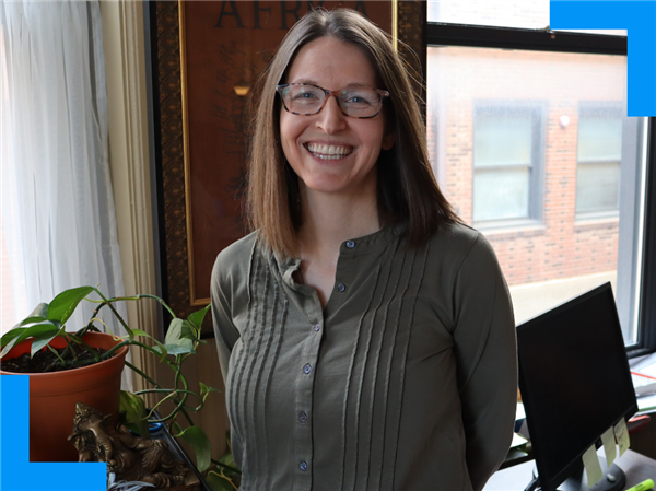 A woman stands in front of a window by a computer, a plant and a bronze Hindu statue.